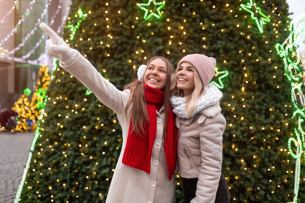 Foto feliz joven en ropa de abrigo apuntando hacia la novia alegre mientras descansa cerca del árbol de navidad decorado con guirnaldas de luz en la calle