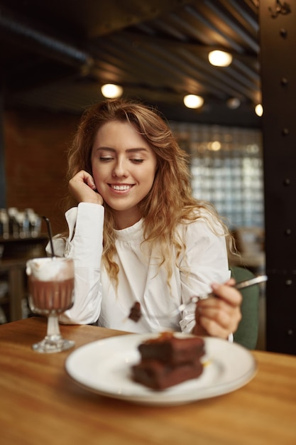 Feliz joven en ropa casual pasar tiempo libre en la acogedora cafetería con pastel de chocolate y café recién hecho