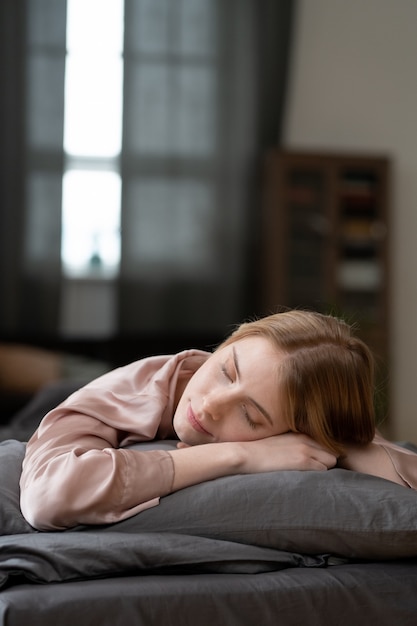 Feliz joven rubia en pijama de seda beige manteniendo la cabeza sobre una almohada suave mientras está acostada en la cama frente a la cámara contra la ventana con cortinas