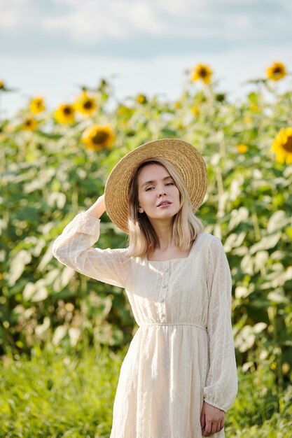 Feliz joven rubia con sombrero y vestido blanco romántico de pie sobre la hierba verde frente a la cámara contra el campo de girasol en un día soleado