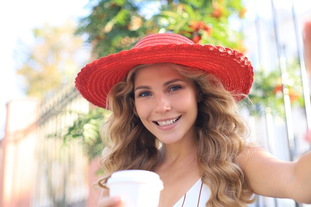 Feliz joven rubia sonriendo ampliamente haciendo autorretrato en su dispositivo al aire libre.