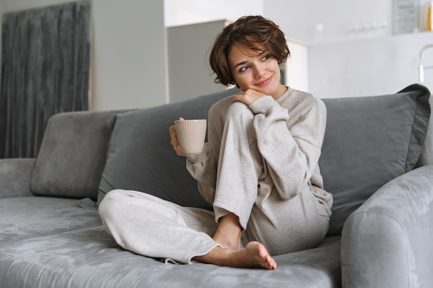 Feliz joven sentada en un sofá en casa, sosteniendo la taza