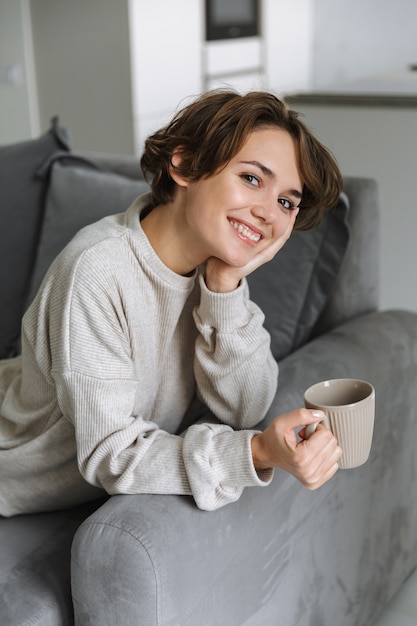 Feliz joven sentada en un sofá en casa, sosteniendo la taza