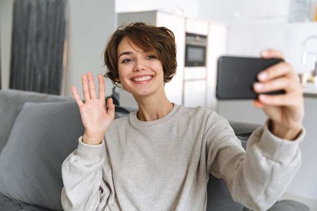 Feliz joven sentada en un sofá en casa, mediante teléfono móvil, haciendo una videollamada