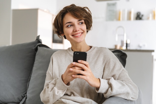 Feliz joven sentada en un sofá en casa, mediante teléfono móvil