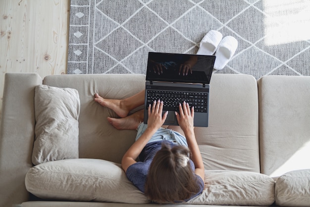 Feliz joven sentada en el sofá y usando la computadora portátil en casa