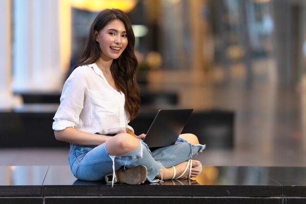 Feliz joven sentada en el suelo con el uso de la computadora portátil en la noche en la ciudad