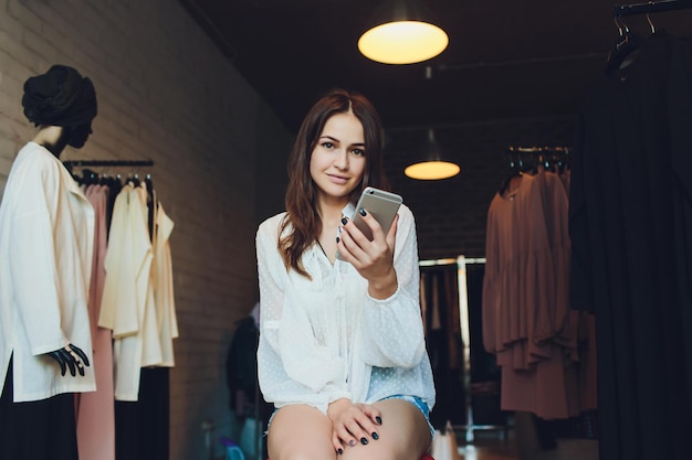 Feliz joven sentada en la tienda de ropa con bolsas de compras y hablando por teléfono móvil