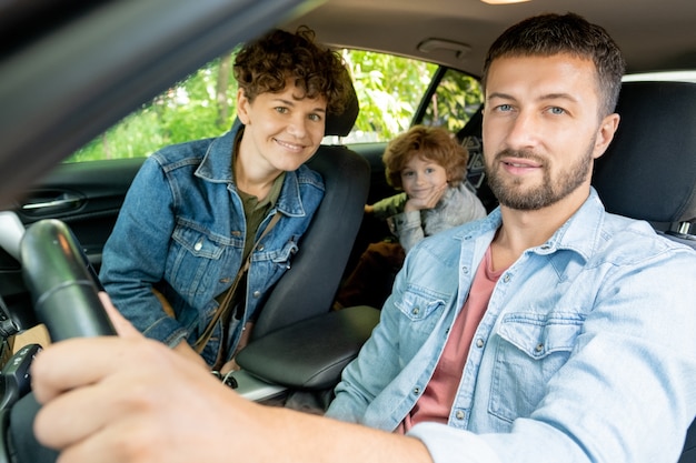 Feliz joven sentado al volante en el coche con una bonita esposa y su lindo hijo en el asiento trasero mirándote
