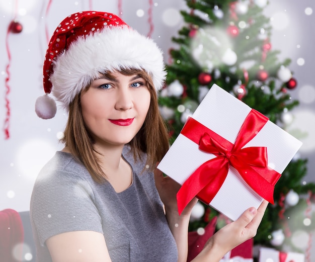 Feliz joven con sombrero de santa con caja de regalo cerca del árbol de navidad decorado