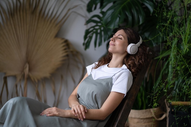 Foto feliz joven soñadora con auriculares descansando rodeada de varias plantas exóticas tropicales