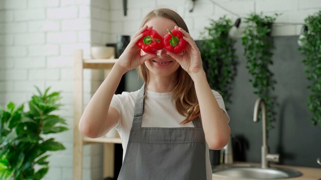 Feliz joven sonríe y se divierte jugando con pimiento rojo frente a sus ojos en la cocina de casa