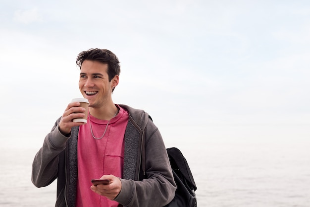 Feliz joven sonriendo con un café y utilizando el concepto de tecnología al aire libre de teléfono móvil