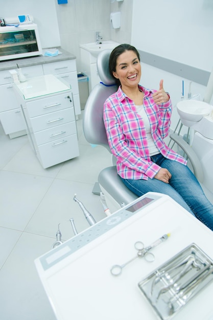 Foto feliz joven sonriendo y levantando su pulgar mientras está sentado en un sillón médico en un consultorio dental moderno