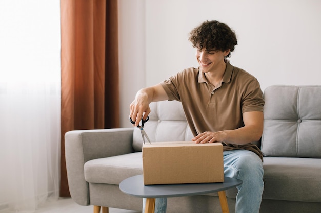 Feliz joven sonriente hombre rizado abriendo caja con productos pedidos regalos en casa en el sofá Comprador en línea cliente masculino abriendo paquete de tienda en línea