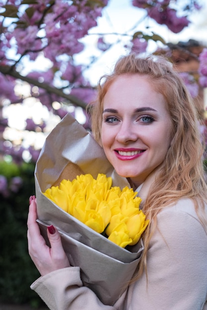 Feliz joven sonriente con ojos azules sosteniendo ramos de tulipanes amarillos árbol en flor en el fondo