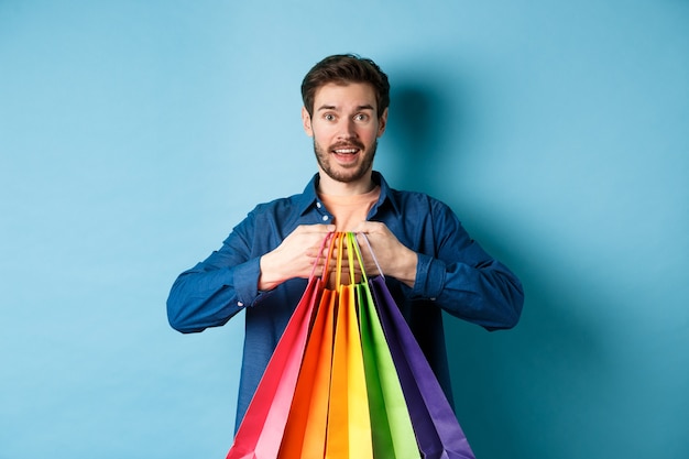 Feliz joven sosteniendo coloridas bolsas de la compra y sonriendo emocionado, comprando regalos, de pie sobre fondo azul.