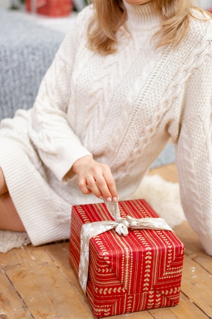 Feliz joven sosteniendo un presente rodeado de cajas de regalo se sienta con las piernas cruzadas en un sofá de camello. Regalo de Navidad en manos de una niña, primer plano.