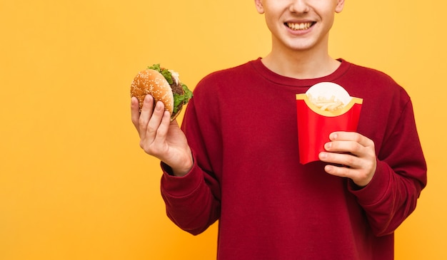 Feliz joven en sudadera roja con hamburguesa y papas fritas en las manos
