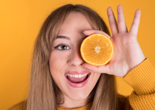 Feliz joven en suéter naranja con naranjas cubriendo un ojo, pared de color amarillo pastel