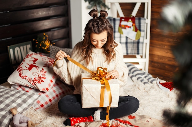 Feliz joven en suéter de punto vintage en la cama abriendo un regalo en Nochebuena
