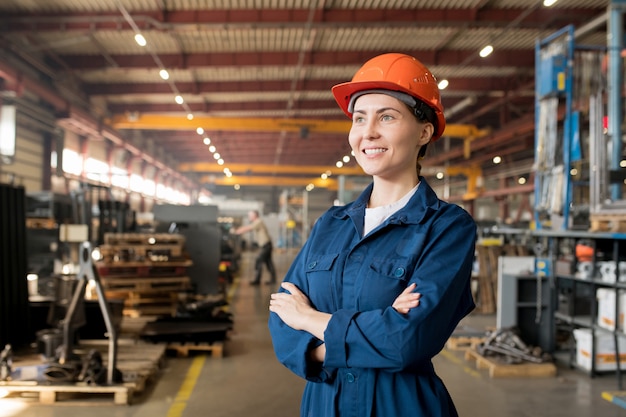 Foto feliz joven técnico en ropa de trabajo azul y casco de pie dentro de un gran taller en la fábrica.