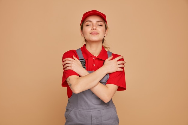 Feliz joven trabajador de la construcción vistiendo uniforme y gorra abrazándose a sí misma con los ojos cerrados