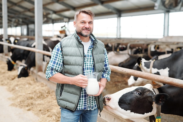 Feliz joven trabajador de la granja lechera sosteniendo una jarra con leche fresca mientras está de pie junto al establo, una de las vacas oliéndole