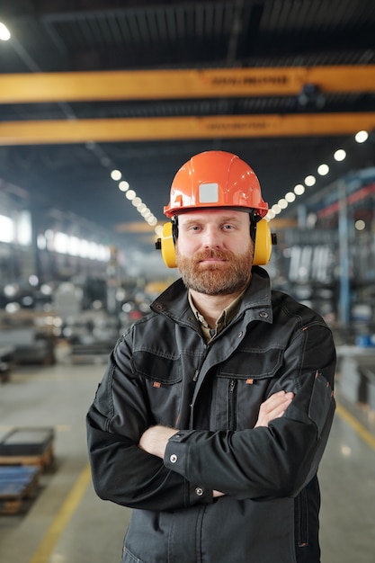 Feliz joven trabajador industrial barbudo en auriculares protectores y casco posando