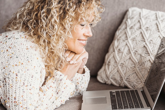 Feliz joven tumbado frente a la computadora portátil en el sofá con las manos juntas en la sala de estar en casa. Empresaria en casual trabajando desde casa. Mujer caucásica, sonriente, mientras, utilizar la computadora portátil