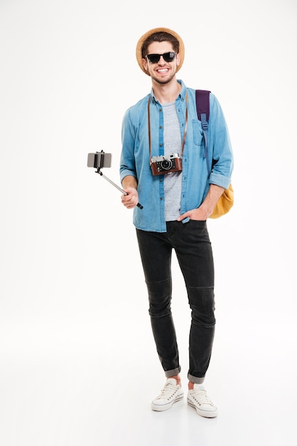 Feliz joven turista sosteniendo mochila, cámara vintage y smartphone en selfie stick sobre fondo blanco.