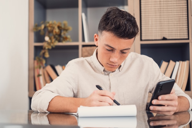 Feliz joven usando su teléfono inteligente en casa navegando por la red haciendo los deberes divirtiéndose solo en el interior. Adolescente masculino con teléfono disfruta de tiempo libre estudiando y trabajando.