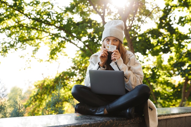 Feliz joven vestida con abrigo y sombrero de otoño sentado al aire libre, usando una computadora portátil, mostrando una tarjeta de crédito de plástico