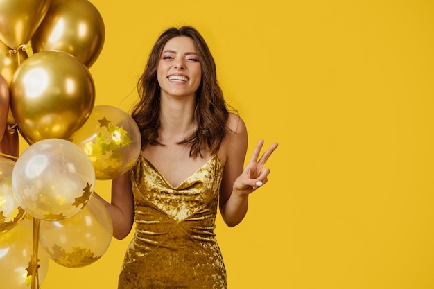 Foto feliz joven vestida sosteniendo globos y mostrando el signo de la paz con la mano aislada en el espacio de copia de fondo amarillo