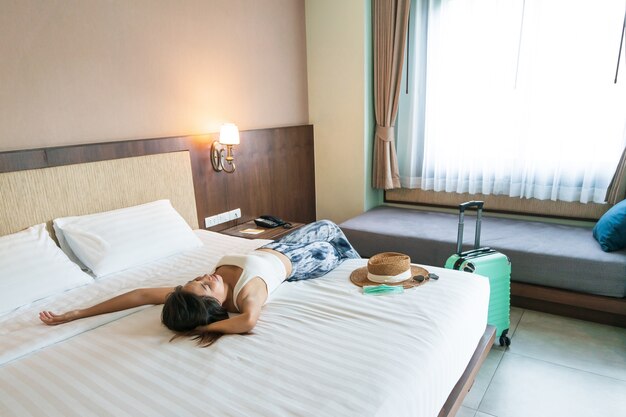 Feliz joven viajero asiático mujer relajarse en la cama con sombrero y máscara médica en la cama en la habitación del hotel