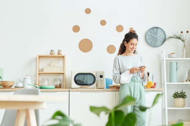 Feliz joven vistiendo elegante traje de pie en su cocina enviando mensajes de texto a alguien que usa su teléfono inteligente