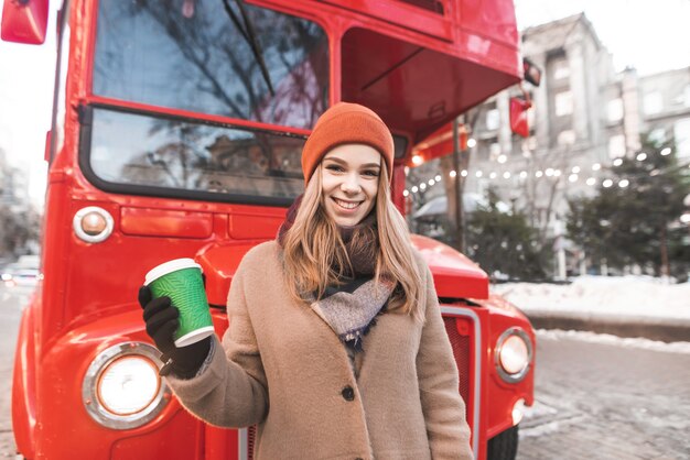 Feliz joven vistiendo una gorra y ropa de abrigo se encuentra en el fondo de un autobús rojo con una taza de café en la mano, mira a la cámara y sonríe