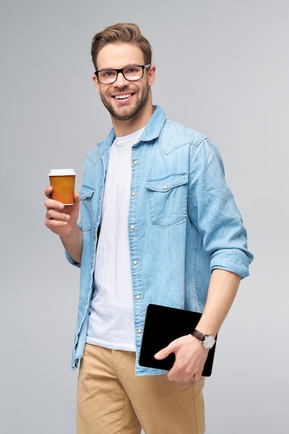 Feliz joven vistiendo jeans camisa de pie y usando tablet pc pad y sosteniendo una taza de café para llevar