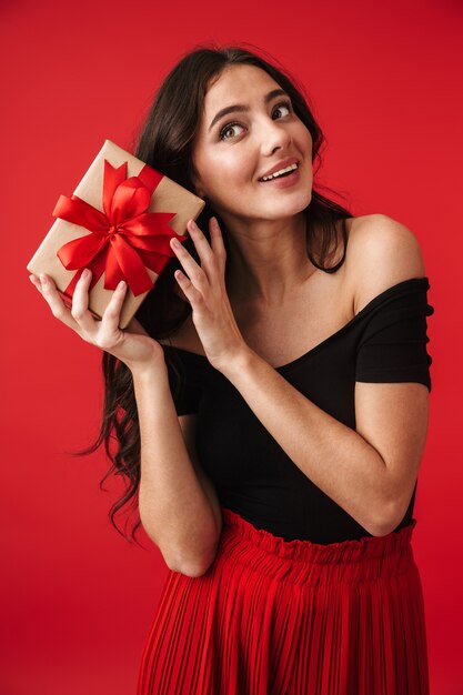 Feliz joven vistiendo un vestido que se encuentran aisladas sobre rojo, sosteniendo una caja de regalo