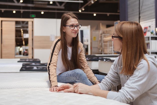 Feliz jovencita hablando con su mamá mientras elige un nuevo colchón ortopédico para comprar