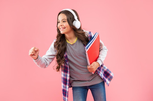 Feliz jovencita con libro usando audífonos sobre fondo rosa, escuela