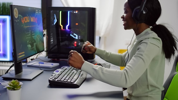 Feliz jugador negro levantando las manos ganando la competencia en línea de tirador espacial hablando con un micrófono profesional en un estudio de juegos