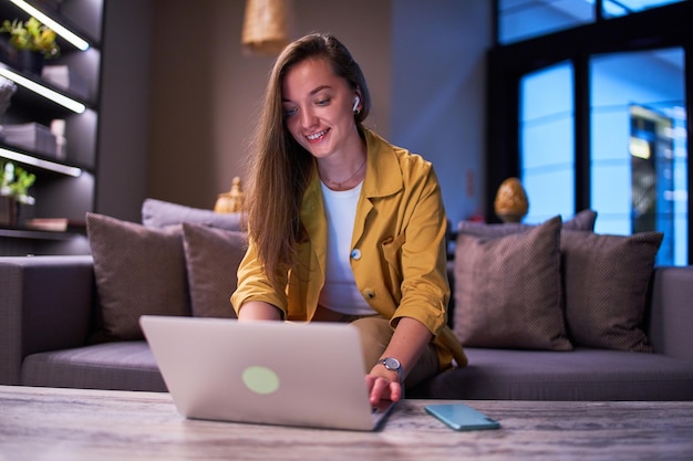 Feliz, linda, alegre, moderna, sonriente, satisfecha, inteligente, joven, casual, mujer de negocios usando auriculares inalámbricos usando una computadora portátil para trabajar en línea en el café