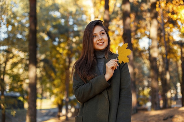 Feliz linda chica con un elegante abrigo gris está caminando en un parque de otoño sosteniendo una hoja amarilla en sus manos. Mujer atractiva.