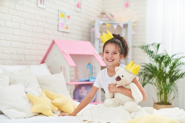 Foto feliz linda chica sentada en la cama en la habitación de sus hijos y abraza el oso de peluche.