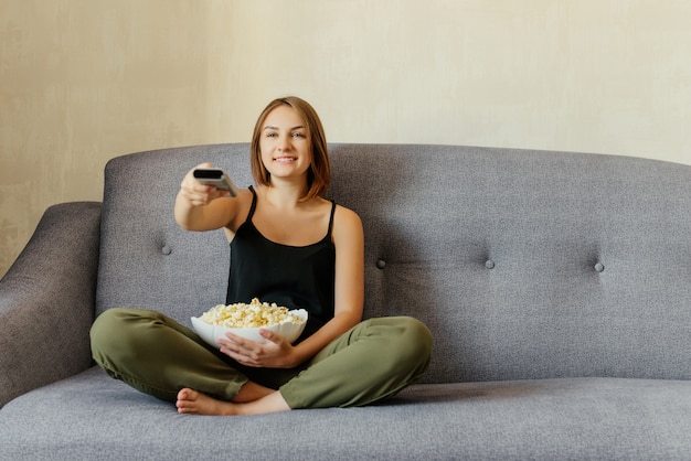 Feliz linda chica sentada en el sofá gris, comiendo palomitas de maíz, disfrutando viendo un televisor. Adentro. Tiempo de relajación.