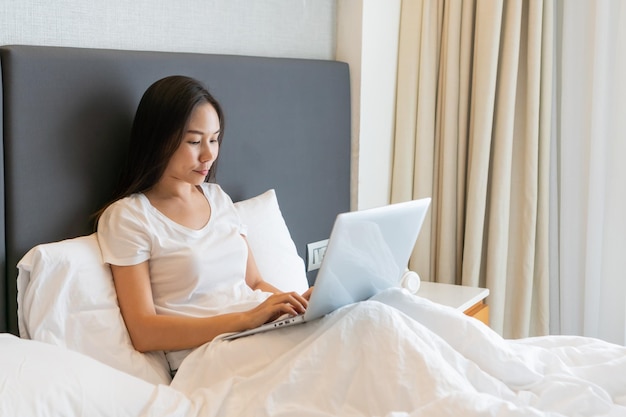 Feliz linda empresária asiática trabalhando em um laptop enquanto está sentado na cama em casa Trabalhe em casa Conceito de tecnologia e estilo de vida Closeup copy spacex9