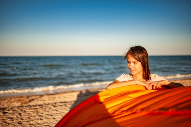 Feliz linda garota com cabelo escuro fica perto de uma tenda brilhante sorrindo na praia do mar azul brilhante