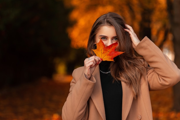 Foto feliz linda garota com casaco elegante cobre o rosto com folhas coloridas de outono no parque outono dourado. férias de outono