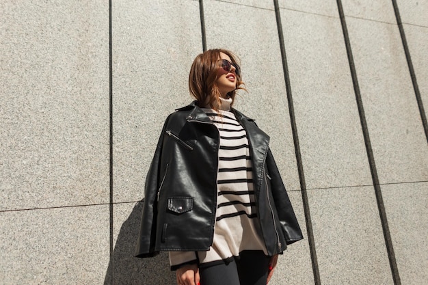 Feliz linda garota de rock urbano de moda jovem com óculos de sol em uma jaqueta de couro preta e um elegante suéter listrado em um dia ensolarado na parede